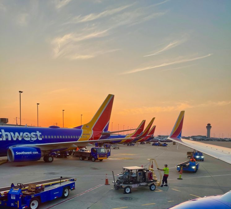 A vibrant scene on the tarmac at sunset features several Southwest Airlines planes lined up in a row, their distinctive red, yellow, and blue tail fins glowing in the warm evening light. Ground crew members and vehicles are actively preparing for operations as the airport's control tower is visible in the distance. The sky is painted with a soft gradient of orange and blue, enhancing the calm yet busy atmosphere.