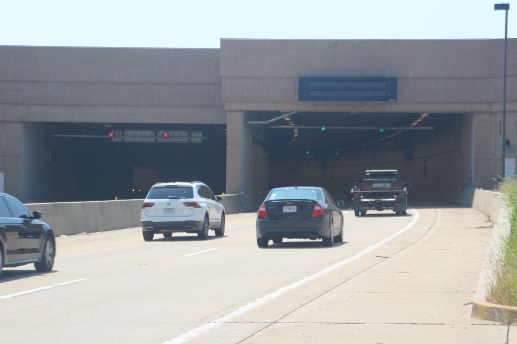 Cars are seen entering a wide concrete tunnel with overhead green arrows indicating open lanes and red Xs marking closed lanes.