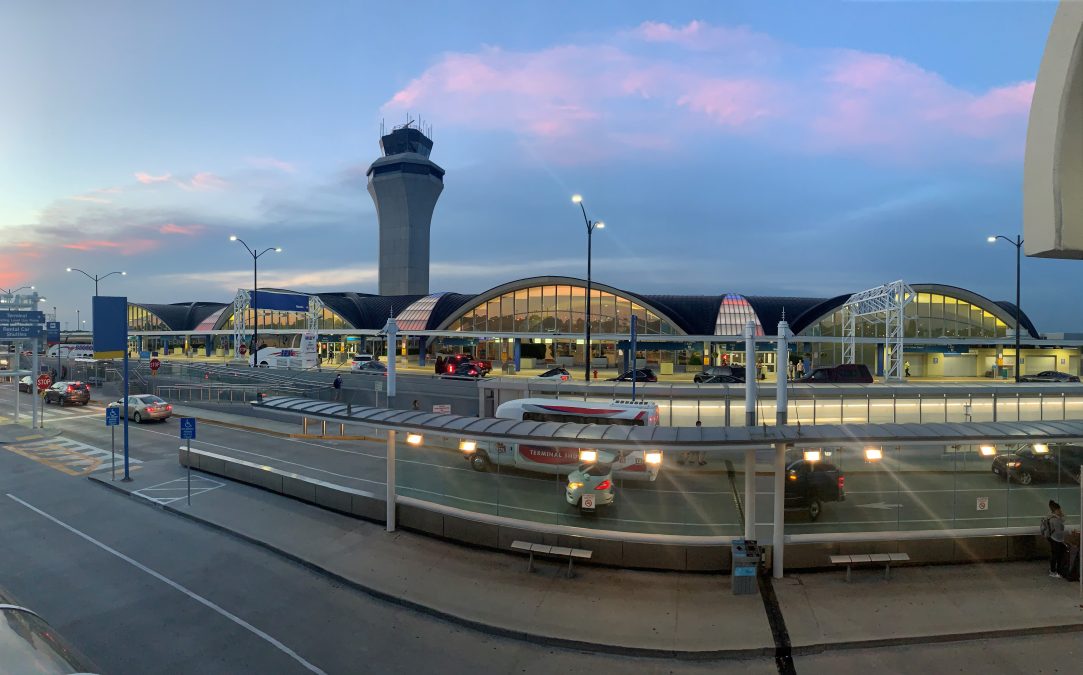 The exterior of Terminal 1,at St. Louis Lambert International Airport glows under the evening sky, with a mix of deep blues and soft pink hues in the clouds. The iconic control tower rises prominently above the curved terminal roof, while cars and shuttles move through the well-lit drop-off and pickup lanes below. The scene captures a calm yet active airport atmosphere at dusk.
