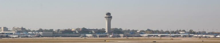 The image shows a panoramic view of St. Louis Lambert International Airport's terminal area and air traffic control tower. The terminal's iconic curved architecture is prominently visible in the center, with aircraft and ground support equipment spread across the tarmac in front of it. The control tower rises centrally, dominating the skyline against a clear, hazy sky.