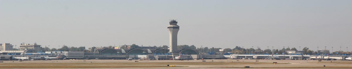 The image shows a panoramic view of St. Louis Lambert International Airport's terminal area and air traffic control tower. The terminal's iconic curved architecture is prominently visible in the center, with aircraft and ground support equipment spread across the tarmac in front of it. The control tower rises centrally, dominating the skyline against a clear, hazy sky.