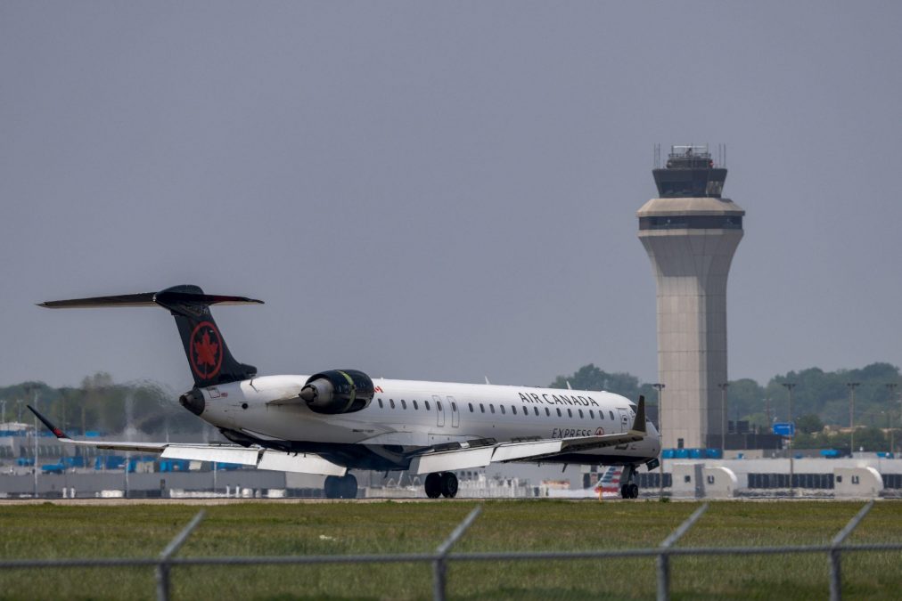 An Air Canada Express plane is positioned on the taxiway at St. Louis Lambert International Airport. The aircraft, with the airline's maple leaf logo prominently displayed on its tail, is seen against the backdrop of the airport's control tower. The scene features grassy areas, fencing in the foreground, and airport terminal infrastructure in the distance.