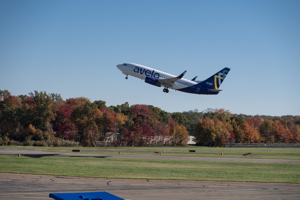 An Avelo Airlines plane is taking off from St. Louis Lambert International Airport, with its landing gear still extended. The aircraft is ascending against a clear blue sky. Below, a grassy field and taxiway are bordered by trees showing vibrant fall colors in shades of red, orange, and yellow.