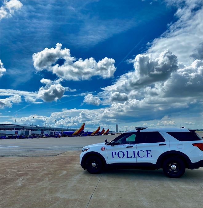 Photo of a police car on a runway with a blue sky and planes lined up in the background