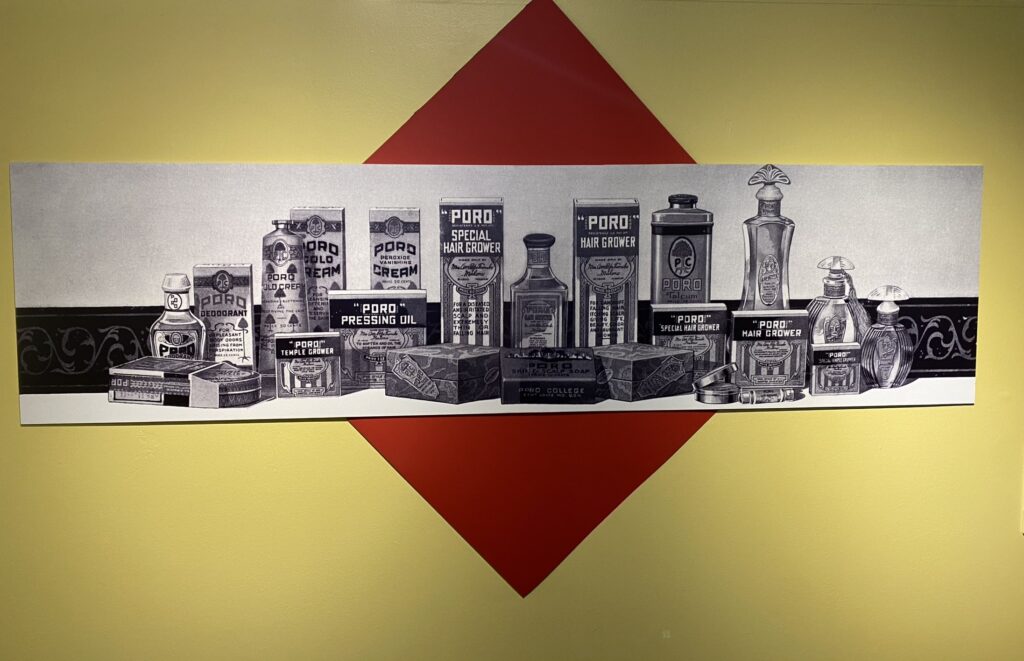 This image is a display showcasing vintage "Poro" beauty and hair care products. The black-and-white artwork highlights items such as Poro Hair Grower, Pressing Oil, Talcum, and Temple Grower, arranged in front of a decorative panel with a diamond-shaped red backdrop. These products are part of the legacy of Annie Malone, a pioneer in African American hair care and cosmetics.