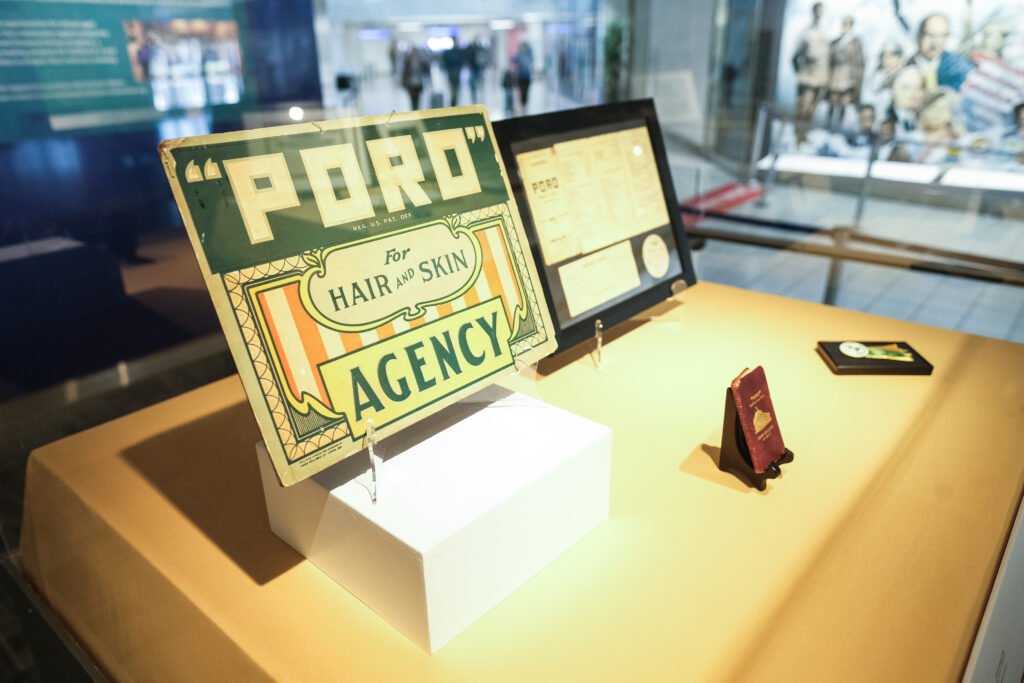 This image showcases a display case featuring memorabilia related to Annie Malone's Poro brand. The centerpiece is a vintage sign reading "Poro for Hair and Skin Agency." Additional items include framed Poro documents, certificates, and a small red booklet.