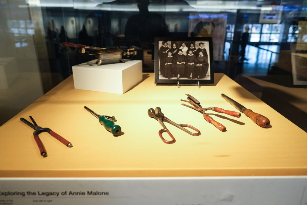 This image features a display case showcasing vintage hair styling tools from the Poro College era, including curling irons, hair crimpers, and other implements with wooden and metal handles. A framed black-and-white photograph of Poro College graduates in their gowns is also part of the exhibit.