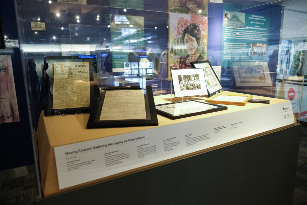 This image showcases a museum exhibit featuring framed documents, photographs, and memorabilia related to Poro College and Annie Malone's business ventures. Notable items include "The Poro Purpose," a publication outlining the mission and vision of Poro, as well as historical certificates, contracts, and a group photograph. A nearby backdrop features artwork and informational panels celebrating her legacy.