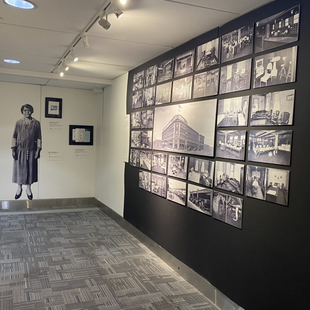 This image features a museum exhibit dedicated to Annie Malone's legacy, with a large wall display of historical photographs showcasing the interior and exterior of Poro College. The images depict various rooms, including classrooms, beauty salons, and offices, illustrating the operations and grandeur of the college. A life-sized cutout of Annie Malone stands to the left, emphasizing her role as a trailblazing entrepreneur in African American beauty and education.