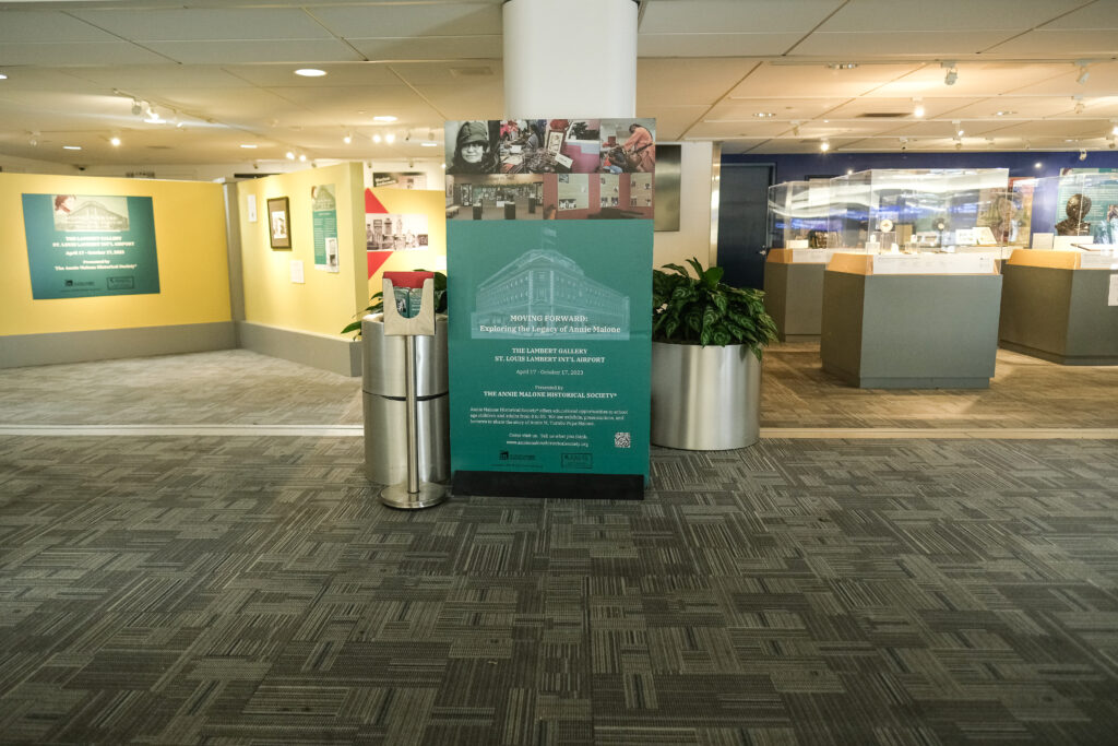 This image showcases the entrance area of the "Moving Forward: Exploring the Legacy of Annie Malone" exhibit at the Lambert Gallery in St. Louis Lambert International Airport. A large informational sign stands prominently in the foreground, providing details about the exhibit's dates, sponsors, and organizers, including the Annie Malone Historical Society. In the background, sections of the exhibit are visible, featuring various display cases, framed artwork, and artifacts related to Annie Malone's groundbreaking work in beauty, education, and entrepreneurship. The bright, open gallery space invites visitors to explore her impactful legacy.
