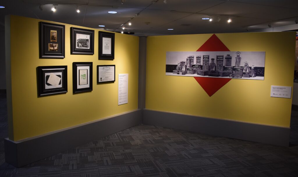 This image showcases a section of the "Moving Forward: Exploring the Legacy of Annie Malone" exhibit. On the left, framed archival materials, including photographs, documents, and a diploma from Poro College, are displayed on a bright yellow wall. To the right, a large black-and-white mural presents an assortment of vintage Poro beauty products arranged against a red diamond-shaped backdrop.