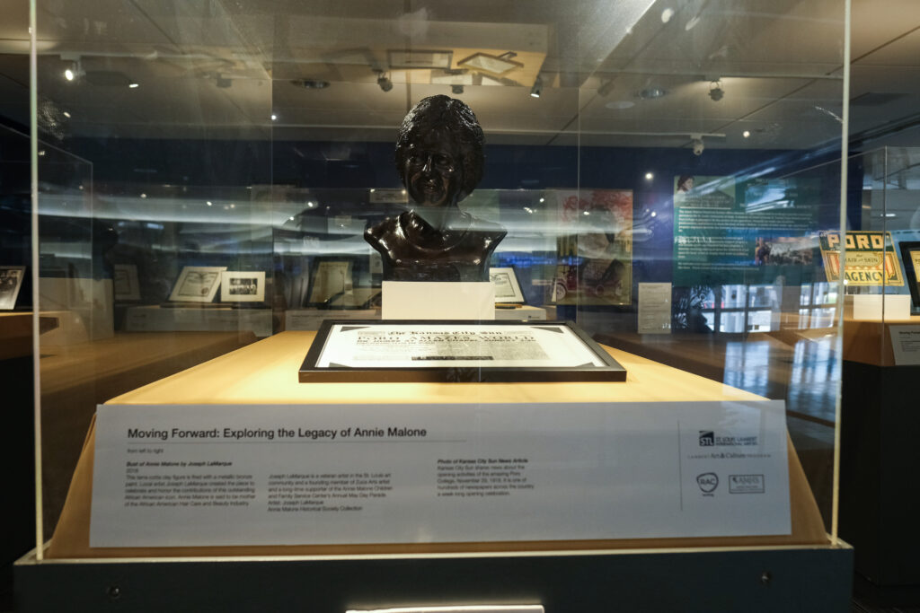 This image shows a museum display featuring a bronze bust of Annie Malone, created by Joseph LaMarque, housed in a glass case.