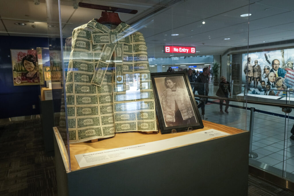 This image showcases a museum display featuring a unique jacket made from one-dollar bills, displayed alongside a framed historical photograph of Annie Malone wearing a similar garment.