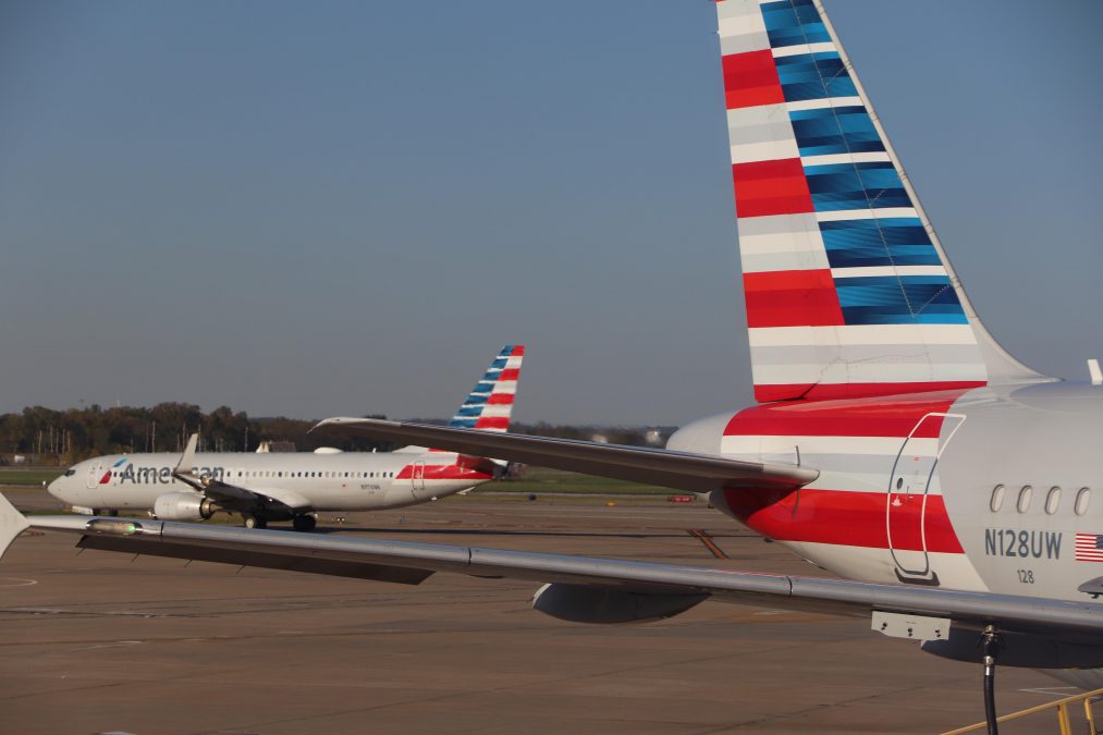 Photo of two American Airlines planes on a runway, facing opposite directions. Both have red, white, and blue tailfins