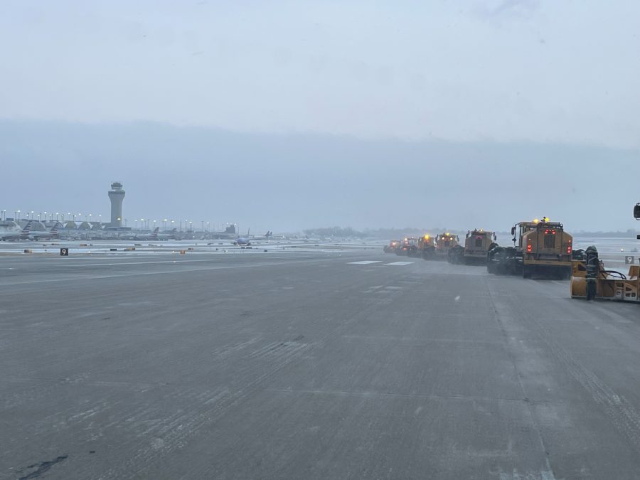 Photo of multiple maintenance vehicles clearing a runway of ice and snow with an airport in the distant background