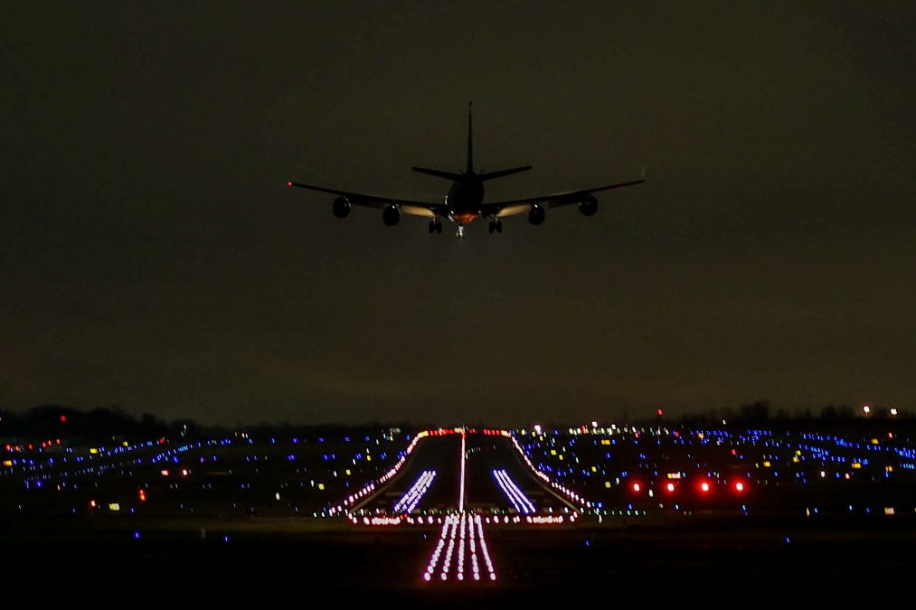Photo of an underlit plane flying away into a night sky with runway lights beneath