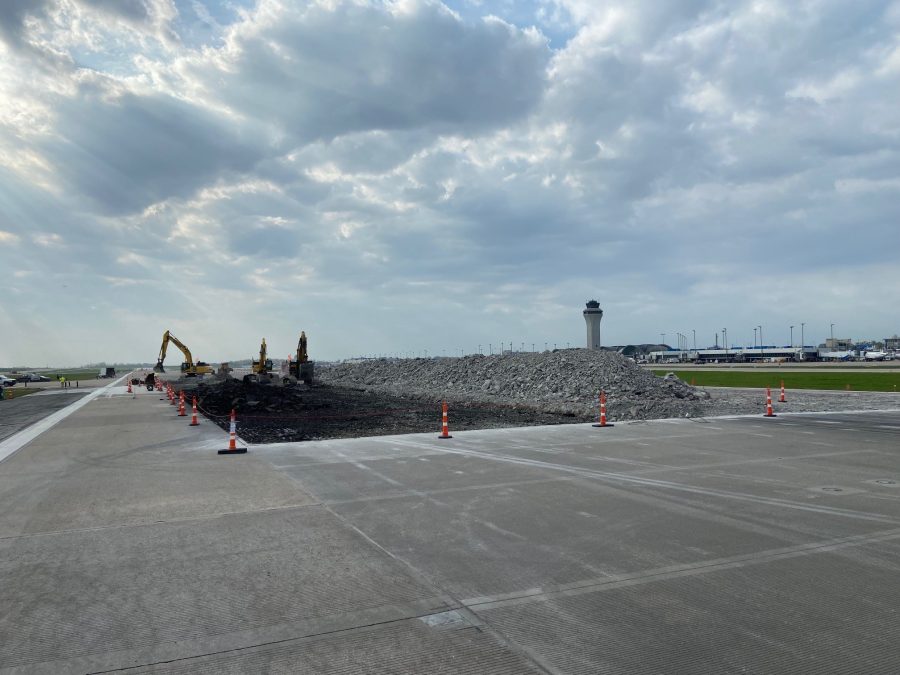Photo of construction on a runway with an airport and airport tower in the distant background