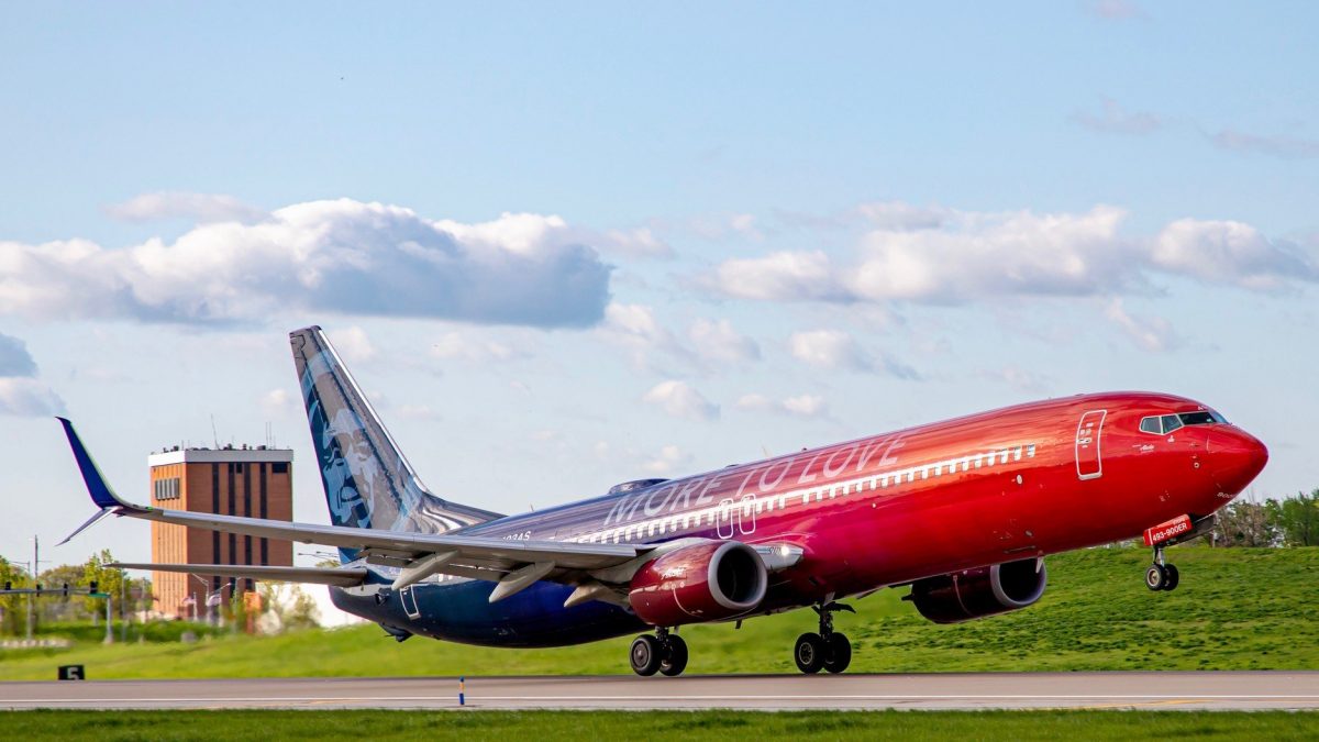 Photo of an Alaska Airlines Boeing 737 that is painted blue and red and taking off of a runway with clouds and a building in the background