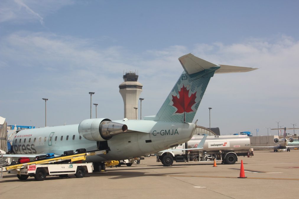 Photo of the back of an Air Canada plane with a red leaf on the tail. The plane appears to be parked at the gate of an airport terminal.