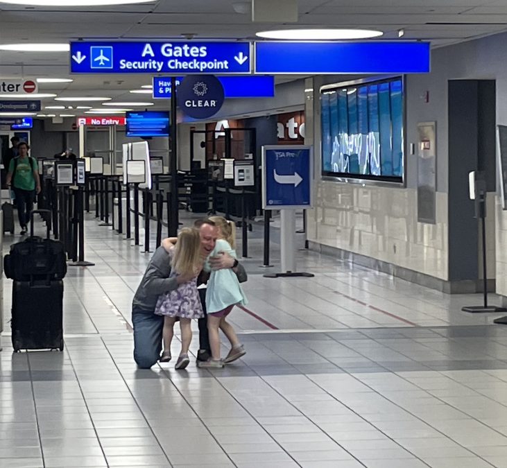 Photo of a man hugging two small children and smiling underneath a sign for an airport gate that says "A Gates. Security Checkpoint"