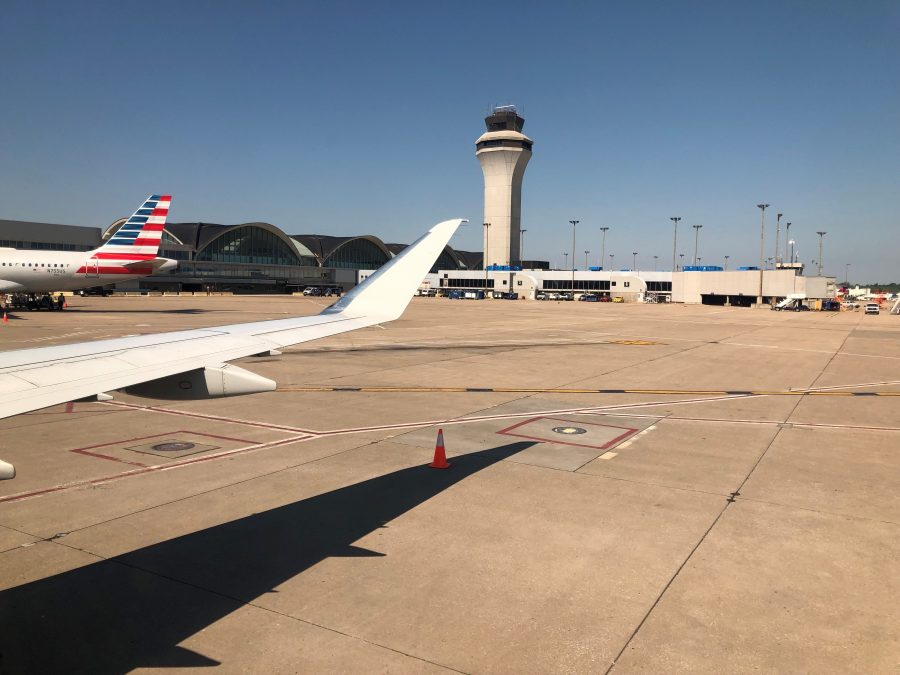Photo of a runway and airport in the background, seemingly taken from the inside of a plane with the plane's wing showing
