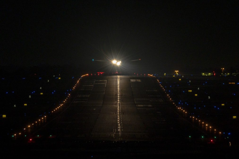 Photo of a lit up runway at night with a plane approaching. The front lights of the plane are bright