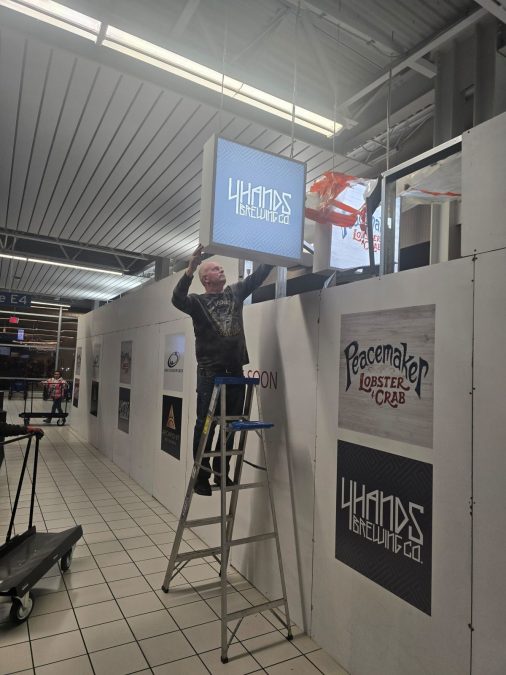 A man stands on a ladder hanging the 4 hands brewing co sign in the airport.