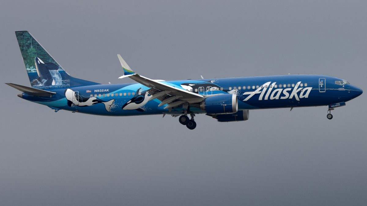 Picture of an Alaskan airlines plane in the sky, decorated in blue with killer whales painted on.