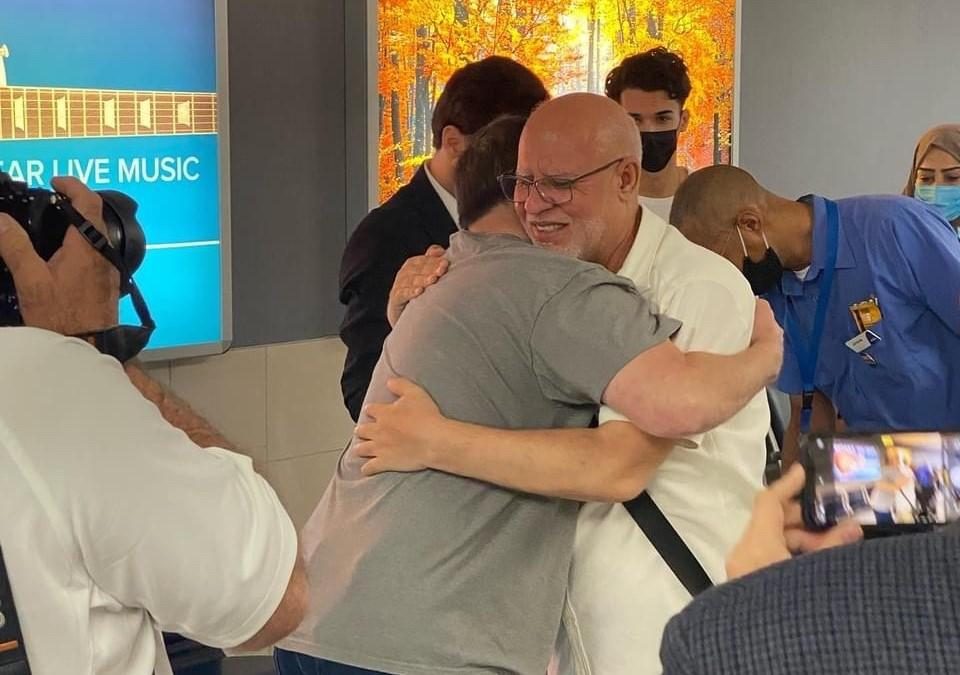Photo of multiple people standing in what appears to be a hallway. Two of the people are hugging and one is smiling