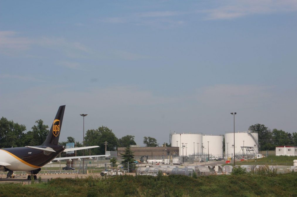 Photo showing a blue sky with the back of a UPS plane visible at the bottom and a fuel farm with white stacks