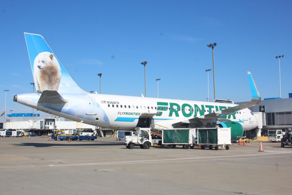Photo of the side of a Frontier Airlines plane on a runway with a sunny sky behind it