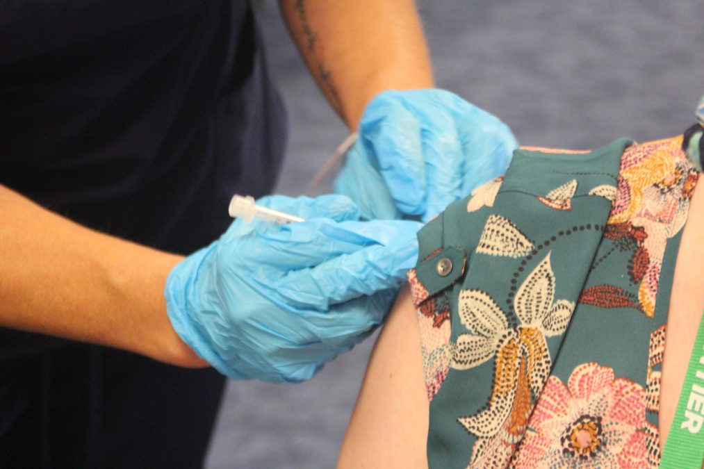 Photo of a person getting a shot in their upper arm from an individual wearing blue gloves