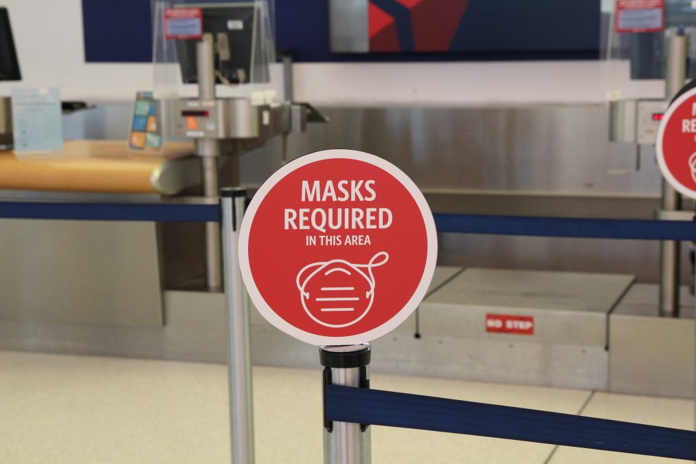 Photo of the inside of an airport terminal with computers in the background and a red sign in the forefront that says "Masks Required In This Area"
