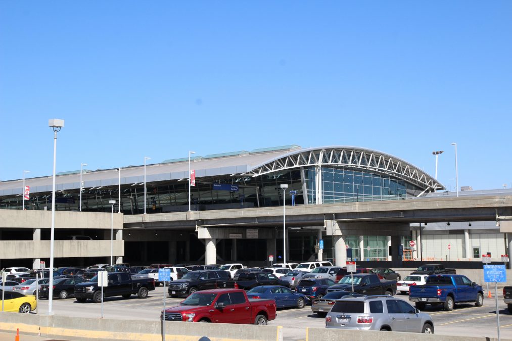 Photo of the outside of an airport terminal with a parking lot containing cars in the foreground