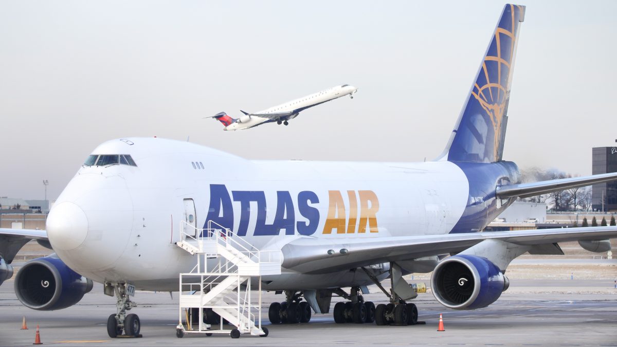 A massive Atlas Air cargo plane dominates the foreground, with its large frame and extended front access stairs emphasizing its size. In the background, a much smaller Delta Airlines aircraft takes off, offering a striking contrast between the two planes.