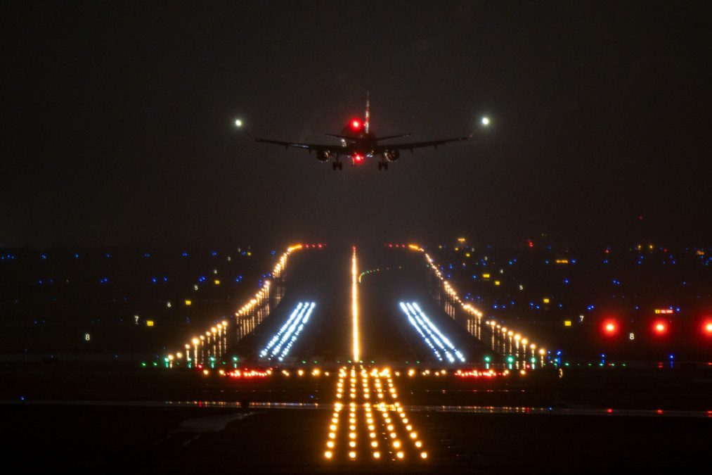 A commercial airplane is approaching a runway for landing at night, with its landing lights shining brightly. The runway is illuminated by rows of white, yellow, and blue lights guiding the aircraft. Red lights and numbered signs are visible on the sides, marking taxiways and runway sections. The dark sky contrasts with the bright lights, emphasizing the dramatic nighttime landing scene.