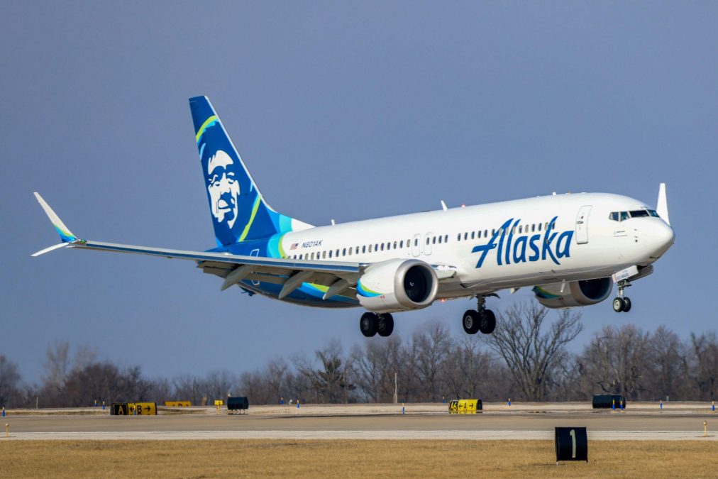 The image captures an Alaska Airlines plane approaching landing at St. Louis Lambert International Airport. The aircraft features the airline's signature white fuselage with the word "Alaska" in bold blue lettering, and a tail design showcasing a stylized portrait of an Alaska Native. The plane is low to the runway with its landing gear extended, set against a clear sky and bare trees in the background.