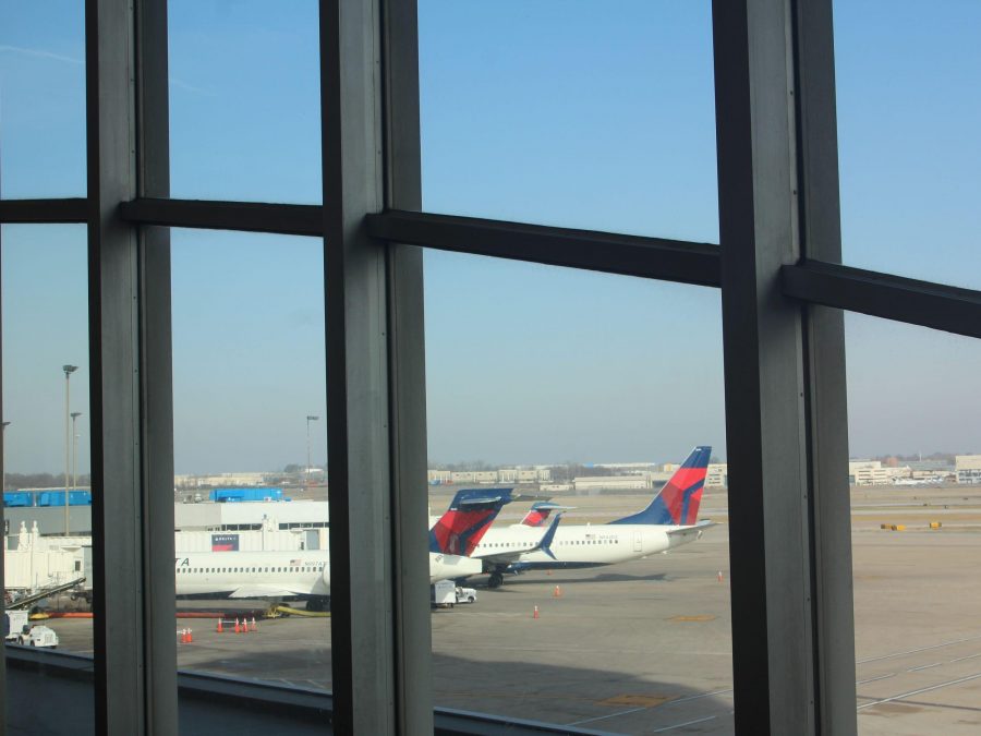 The image shows a view from inside St. Louis Lambert International Airport, looking out through large glass windows onto the tarmac. Delta Airlines planes are parked near the terminal gates, with their red, blue, and white tails visible. The sky is clear, and the airport's operations are seen in the background with other structures and equipment.