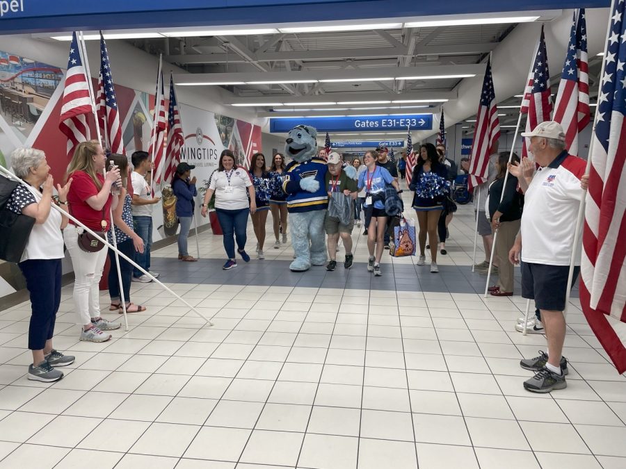 A heartwarming moment unfolds at St. Louis Lambert International Airport as a group of veterans and their escorts walk through a corridor lined with American flags. They are greeted by enthusiastic cheers from supporters and Louie, the St. Louis Blues mascot, adding a festive spirit to the welcome.