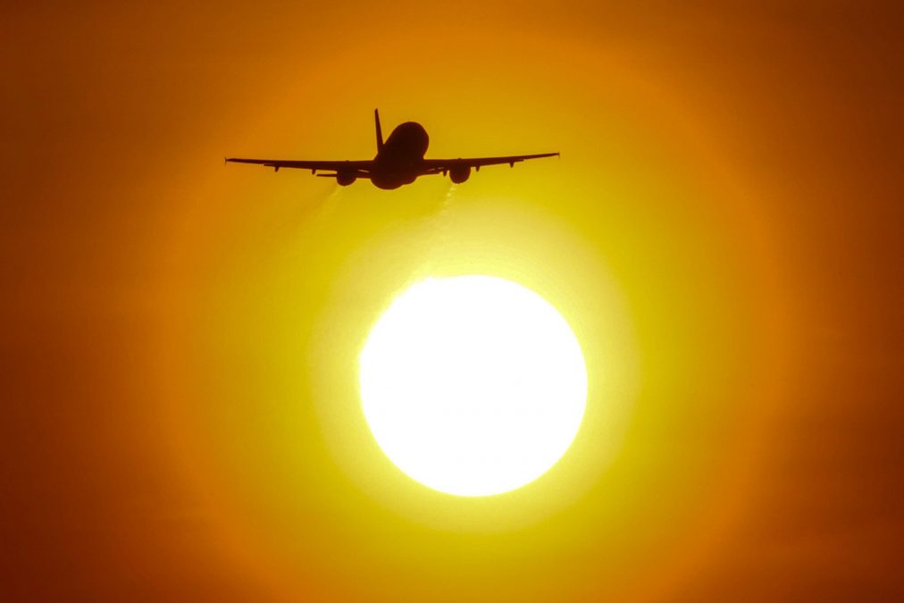 A dramatic silhouette of a plane takes flight directly in front of a setting sun, creating a breathtaking image bathed in warm orange and yellow hues. The intense glow of the sun backlights the aircraft, highlighting its wings and tail.