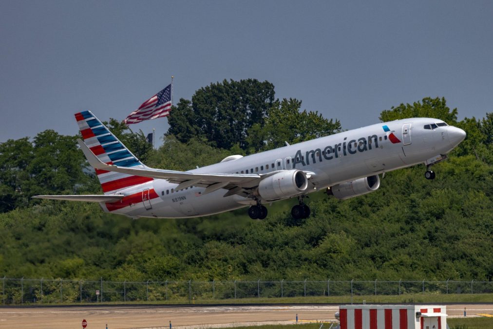 An American Airlines plane lifts off from the runway with an American flag prominently flying in the background. The aircraft's red, white, and blue tail design echoes the patriotic theme, framed by lush green foliage.