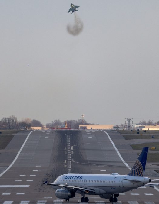 A United Airlines plane waits on the taxiway near a runway, while a F-15 fighter jet performs a near-vertical takeoff in the background, leaving a trail of smoke. The sky is clear, providing an unobstructed view of the runway and surrounding airport area.