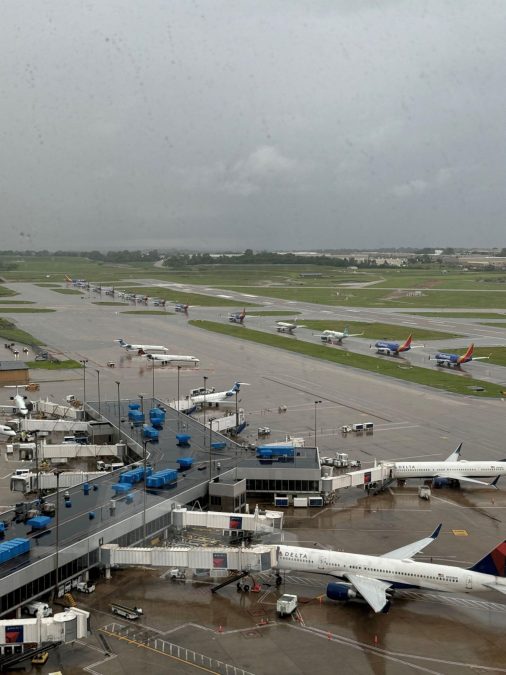 A rainy day view of an airport shows several planes lined up on taxiways and parked at gates, including Delta and Southwest Airlines aircraft. Ground support vehicles and jet bridges are positioned around the terminal, while wet runways and tarmacs reflect the overcast sky. The scene highlights the airport's ongoing operations despite the inclement weather.