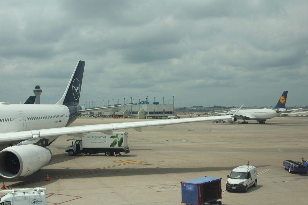 A Lufthansa aircraft sits on the tarmac near a jet bridge, with a wide view of the airport terminal and another Lufthansa plane in the distance. Ground support vehicles, including catering trucks and luggage carts, are stationed near the aircraft. The cloudy sky creates a subdued atmosphere, contrasting with the activity on the ground.