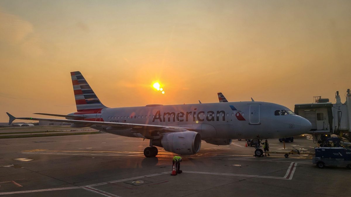 An American Airlines plane is parked at the gate during sunrise, with the sun casting a golden glow across the scene. The aircraft's reflective surface glistens under the warm light as ground crew members work near the plane.