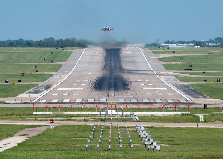 A distant view down an airport runway captures a plane taking off, leaving heat waves and vapor trails in its wake. The runway is lined with a series of evenly spaced approach lights in the foreground and flanked by green fields and taxiway markers. The clear blue sky enhances the expansive perspective and the dynamic sense of movement as the aircraft climbs.