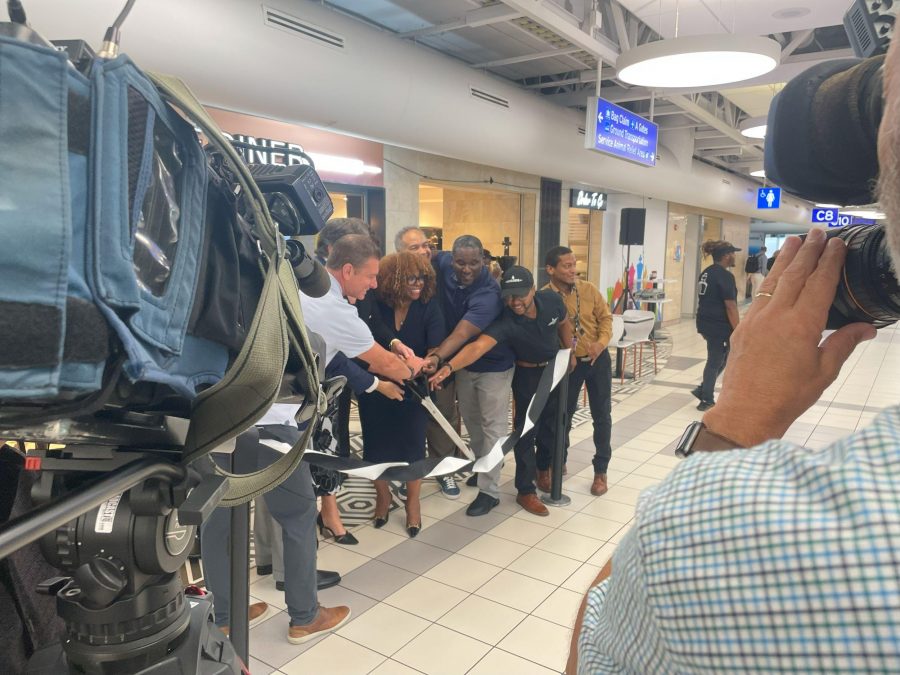 A ribbon-cutting ceremony takes place at STL's Concourse C, marking the opening of a new establishment. Several individuals, including community leaders and business representatives, cut the ribbon together while cameras capture the event. The area is bustling with activity, with a sign for Gate C8 visible in the background.