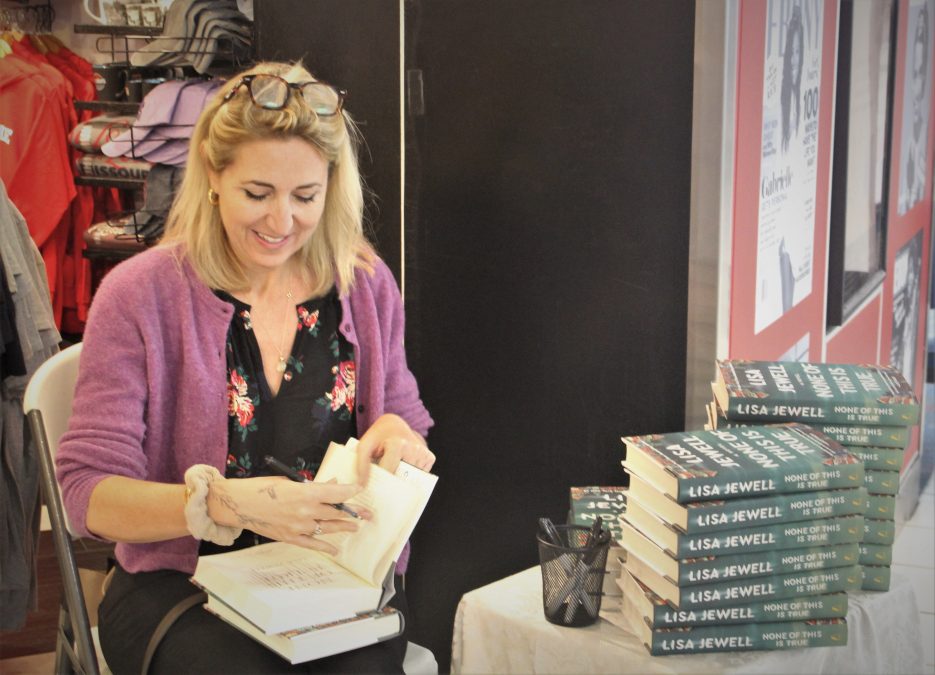 Author Lisa Jewell signs a copy of her book "None of This Is True" at Ebony News in St. Louis Lambert Airport's Concourse C. She sits at a table with a large stack of her books, wearing a purple cardigan, fully engaged in the book signing event. The surrounding display includes posters and airport store merchandise.