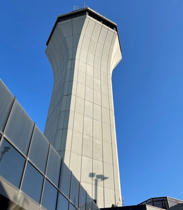 A striking view of the air traffic control tower at St. Louis Lambert International Airport, standing tall against a vivid blue sky. The sunlight casts sharp contrasts on the tower's curved concrete exterior, emphasizing its structural form. Reflections from the nearby glass facade add a modern touch to the composition.