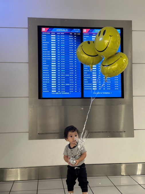 A young child stands in front of a large flight information display screen at an airport, holding three yellow smiley face balloons. The screen behind shows both arrivals and departures information for Terminal 1.